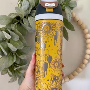 May include: A yellow stainless steel water bottle with a black lid and a silver design of sunflowers, bees, and honeycomb. The lid has a brown tab with the text "Lift to Unlock".