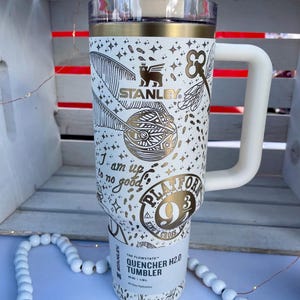 Puede incluir: Un vaso Stanley Quencher H2.0 blanco con detalles dorados y un asa blanca. El vaso presenta una temática de Harry Potter con ilustraciones de una snitch dorada, llaves y el logo de la plataforma 9 3/4. También es visible el texto "I am up to no good".
