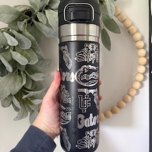 May include: Black insulated travel mug with a silver lid and accents. The mug features etched designs of alligators, the words "Swamp Life," and "Gators." The mug is held by a person.