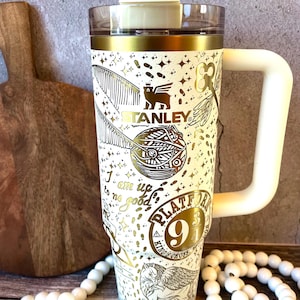 May include: White and gold Stanley tumbler with a handle, featuring a design with golden snitch, Platform 9 3/4, and other Harry Potter-themed graphics. The tumbler has a white handle and a clear lid with a straw.
