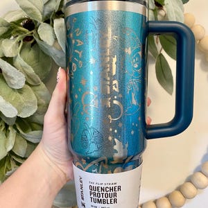 May include: A teal and blue Stanley Quencher Protour Tumbler with a handle. The tumbler has a decorative design and the text "NATION". It holds 887 ml and features "All Day Hydration".