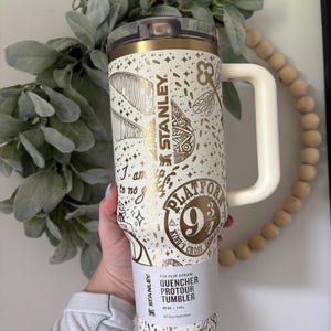 Puede incluir: Un vaso Stanley Quencher Protour blanco y dorado con asa. El vaso presenta ilustraciones doradas de una pluma, una llave y estrellas. El texto en el vaso incluye "Platform 9 3/4" y "All Day Hydration."