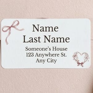 May include: A white rectangular label with rounded corners, printed with the text "Name Last Name" and an address. The design includes a bow and a heart-shaped motif, both in a soft pink colour. The background is a pale pink.