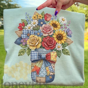 May include: A pale blue sweatshirt with a vibrant floral design. The design showcases sunflowers, roses, daisies, and other flowers in a mason jar. The jar is adorned with patchwork fabric and a blue bow. The word "HoneyPix" is at the bottom.