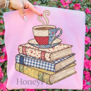 May include: A light pink sweatshirt featuring a graphic of a teacup and saucer atop a stack of books. The teacup and books have a patchwork design with floral, polka dot, and gingham patterns. The word "Honey" is visible at the bottom.