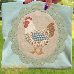 May include: A light blue fabric item featuring a circular appliqué of a chicken. The chicken has a red comb, white neck, and a body with floral and blue gingham patterns. The appliqué is framed by a scalloped edge with small floral details.