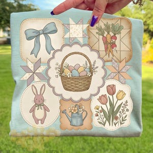 May include: A pale blue t-shirt with various pastel illustrations. Designs include a bow, a basket of eggs, a bunny, carrots, tulips, and a watering can. The shirt has a HoneyPix logo at the bottom.