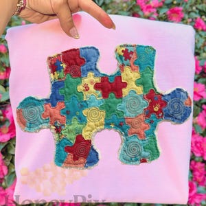 Könnte beinhalten: Ein hellrosa Sweatshirt mit einem großen, farbenfrohen Puzzle-Applikation. Das Puzzleteil besteht aus kleineren, gestickten Teilen in verschiedenen Farben, darunter Rot, Blau, Grün und Gelb. Das Design beinhaltet Handabdrücke und Wirbel.