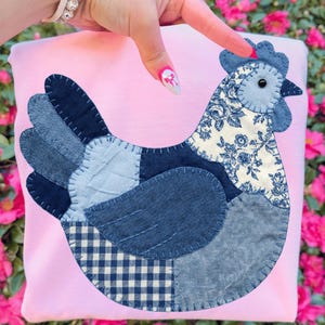Peut inclure: Un article en tissu rose avec un motif de poulet en patchwork. Le poulet est composé de divers patchs en denim bleu, dont un imprimé floral et un motif vichy. Le poulet a une crête et un bec bleus, et un œil noir.