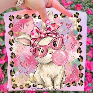 May include: Pink t-shirt featuring a cartoon bunny wearing pink sunglasses and a bow, blowing a bubble. The design includes hearts, gold accents, and a leopard print border. The bunny is white with pink accents.
