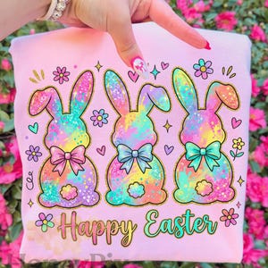 May include: Pink Easter-themed sweatshirt featuring three colorful bunny illustrations with bowties and gold outlines. The words "Happy Easter" are written in a decorative font. The design includes flowers, hearts, and stars.