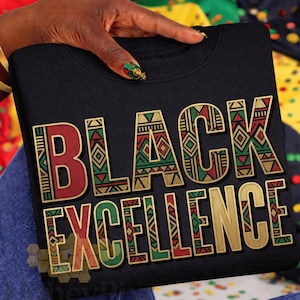 May include: Black crew neck sweatshirt with the words "BLACK EXCELLENCE" in gold, red, and green geometric lettering. The sweatshirt is held by a person with gold bracelets and colorful nail art. The background is a mix of colorful fabrics.