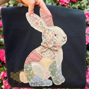 Op de afbeelding: Een zwart stoffen item met een patchwork konijn ontwerp. Het konijn heeft een crèmekleurig lichaam, roze oren en een bloemenstrik. Het patchwork bevat roze, blauwe en gele secties. Het item wordt door een hand omhoog gehouden.