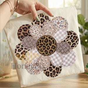 Op de afbeelding: Een beige vierkant kussen met een groot bloemmotief. De bloemblaadjes zijn gemaakt van verschillende stoffen met patronen, waaronder luipaardprint, bloemen en ruitjes. Het midden van de bloem is luipaardprint.