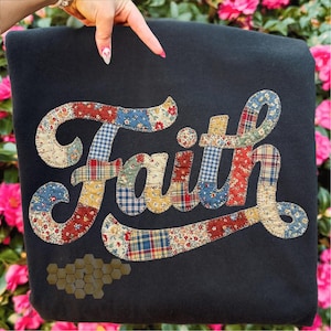 Puede incluir: Una manta negra con la palabra "Faith" aplicada en un diseño de patchwork. Las letras están compuestas por varias telas florales y a cuadros en rojo, azul y amarillo. Un grupo de formas hexagonales marrones está en la parte inferior.