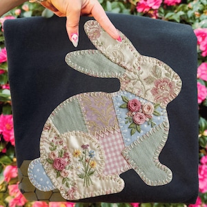 May include: A black fabric item with a patchwork bunny design. The bunny is made from various patterned fabrics, including floral and gingham, with embroidered details and small rose embellishments. The item is held by a hand with pink nail polish.