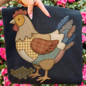 Puede incluir: Una manta azul oscuro con un diseño de gallina de retazos. La gallina está compuesta por varios patrones y colores de tela, incluyendo cuadros, espiga y tonos lisos. La manta tiene una estética rústica y campestre.