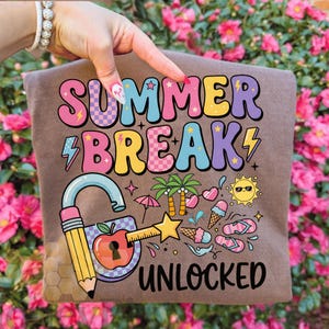May include: A brown t-shirt featuring the text "SUMMER BREAK UNLOCKED" in a vibrant, retro font. The design incorporates a padlock with a pencil key, an apple, and summer icons such as ice cream, sunglasses, and palm trees.
