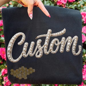 Op de afbeelding: Een donkergrijs stoffen item met het woord "Custom" gespeld in grote, witte parels. Een gouden honingraatpatroon bevindt zich in de linker benedenhoek. Het item wordt omhoog gehouden door een hand met roze en witte nail art.