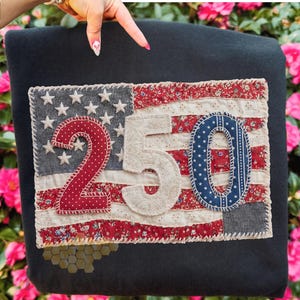 May include: A patriotic textile piece featuring the American flag design with the numbers "250" appliqued on it. The flag incorporates red and white stripes, a blue field with white stars, and the numbers are in red, cream, and blue. The background is a dark gray.