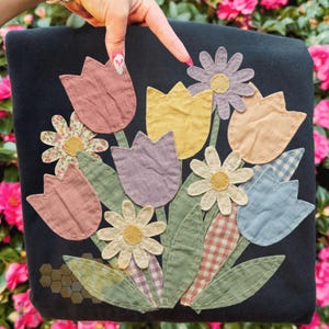 Peut inclure: Un objet en tissu noir avec un motif floral appliqué. L'appliqué comprend des tulipes, des marguerites et des feuilles dans des tons pastel de rose, jaune et bleu. Les fleurs sont cousues sur le tissu, créant un effet texturé.