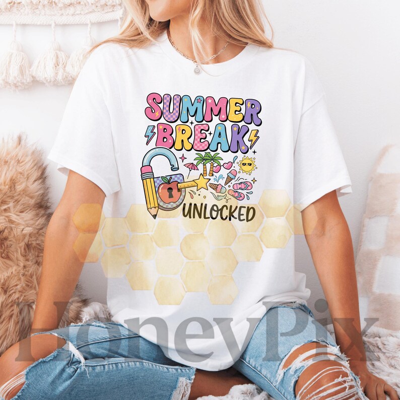 May include: White t-shirt featuring the text "SUMMER BREAK UNLOCKED" in a colourful design. The graphic includes a pencil-shaped lock, palm trees, ice cream, and other summer icons. The shirt is worn by a person wearing blue jeans.