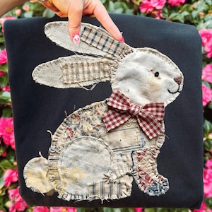 May include: A black pillow featuring a patchwork bunny design. The bunny has a white face, ears with plaid accents, and a red and white checkered bow tie. The bunny is made of various fabric patterns.