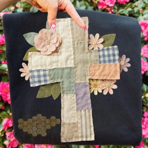 Könnte beinhalten: Ein dunkelblaues Zierkissen mit einem Patchwork-Kreuz-Design. Das Kreuz besteht aus verschiedenen pastellfarbenen Stoffen, darunter Vichy- und Streifenmuster. Das Kissen ist mit Stoffblumen und Blättern verziert und schafft eine rustikale, handgefertigte Ästhetik.