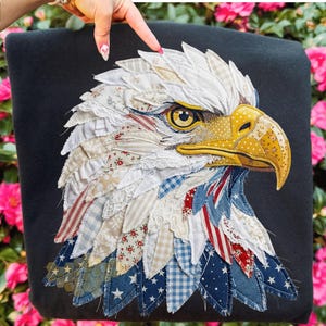 May include: A black textile featuring a detailed appliqué of an eagle's head. The eagle's feathers are crafted from various patterned fabrics in white, red, blue, and beige. The eagle has a yellow beak and eye, with intricate stitching details.