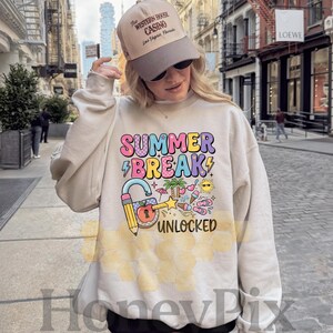 May include: A cream-coloured sweatshirt featuring the words "SUMMER BREAK" in colourful bubble letters, a lightning bolt, and a padlock with a pencil. The design includes summer-themed icons. A tan baseball cap is worn.