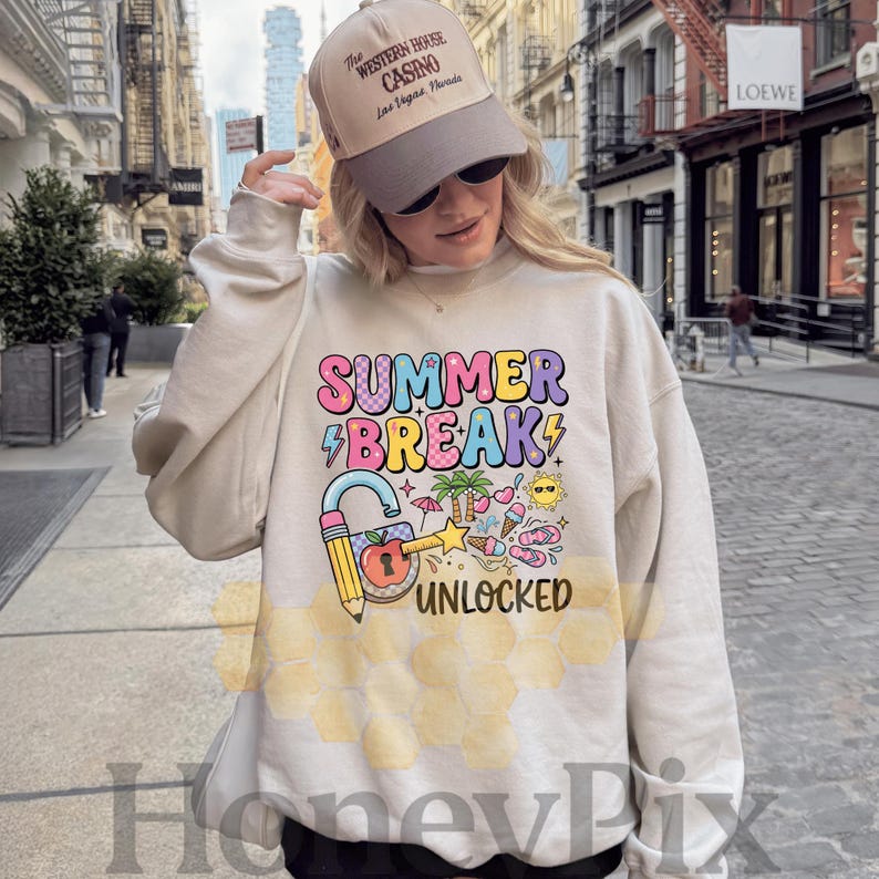 May include: A cream-coloured sweatshirt featuring the words "SUMMER BREAK" in colourful bubble letters, a lightning bolt, and a padlock with a pencil. The design includes summer-themed icons. A tan baseball cap is worn.