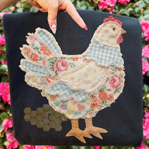 Könnte beinhalten: Ein dunkelblaues Stoffobjekt mit einem Patchwork-Hühnerdesign. Das Huhn hat einen roten Kamm, einen hellblau-karierten Kragen und einen Körper aus Stoffen mit Blumen- und Herzmustern. Ein Wabenmuster ist sichtbar.