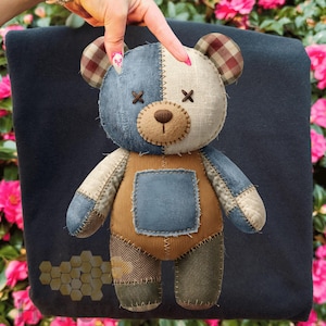 May include: A handmade teddy bear with a patchwork design. The bear features denim, tan, and cream-colored fabric, with brown plaid ears and stitched details. It has a blue square patch on its overalls and is set against a dark blue background.
