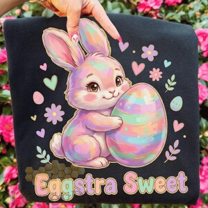 May include: Black sweatshirt featuring a pastel-colored Easter bunny holding a large, decorated egg. The design includes hearts, flowers, and the text "Eggstra Sweet" in a gold-colored font. The overall theme is Easter and springtime.