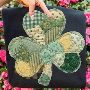 Peut inclure: Un coussin noir en forme de trèfle à quatre feuilles avec un motif patchwork. Le trèfle est composé de divers tissus verts et jaunes avec des motifs floraux, rayés et à pois. Thème de la Saint-Patrick.