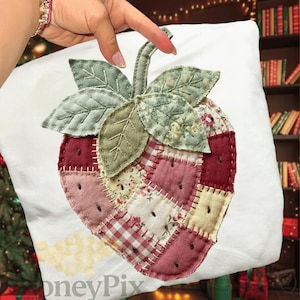 Puede incluir: Un cojín blanco con un gran diseño de fresa aplicado. La fresa está hecha de tela de retazos en tonos rojos, rosas y crema, con hojas y tallo verdes. El diseño tiene una estética casera y acolchada.