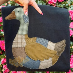 May include: A black throw blanket featuring a patchwork duck design. The duck has a brown beak, a green and white checkered neck, and a body made of various fabric patterns. The wing is tan, and the blanket is held by a hand.