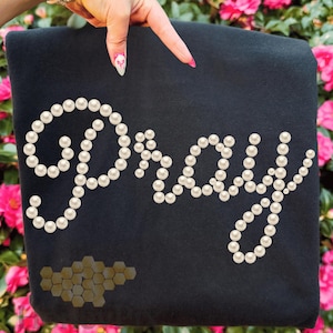 Puede incluir: Tela negra con la palabra "Pray" escrita con perlas blancas. Un grupo de formas de panal doradas se encuentra en la esquina inferior izquierda. La tela está sostenida por una mano con manicura rosa y blanca.
