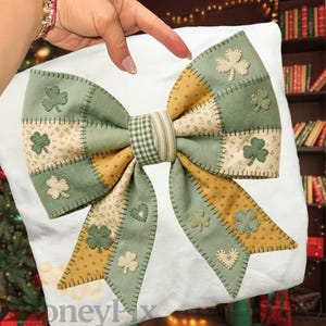 Può includere: Un fiocco decorativo in tessuto patchwork verde e giallo, con applicazioni di trifogli e cuori. Il fiocco è centrato su uno sfondo bianco, creando un aspetto festoso e artigianale, perfetto per San Patrizio.