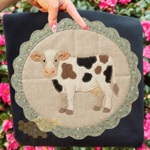 May include: A black cushion featuring a circular appliqué of a cow. The cow has a beige body with brown and black patches. The appliqué is surrounded by a scalloped edge with a floral pattern. The cushion is held by a hand.