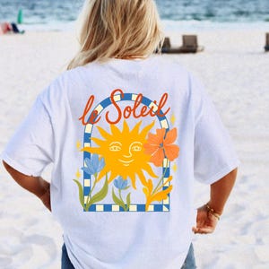 Puede incluir: Camiseta blanca con un gráfico de un sol sonriente y el texto francés "le Soleil" en rojo. El sol es amarillo, con detalles florales naranjas y azules. La camiseta tiene un corte holgado y oversize.