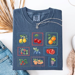 K&ouml;nnte beinhalten: Ein blaues Comfort Colors T-Shirt mit einem Raster von Obstillustrationen in Briefmarkenrahmen. Die Fr&uuml;chte umfassen Zitronen, Kirschen, Orangen, Brombeeren, Blaubeeren, Granat&auml;pfel, Erdbeeren, Pfirsiche und Trauben. Eine silberne Halskette liegt auf dem Shirt.
