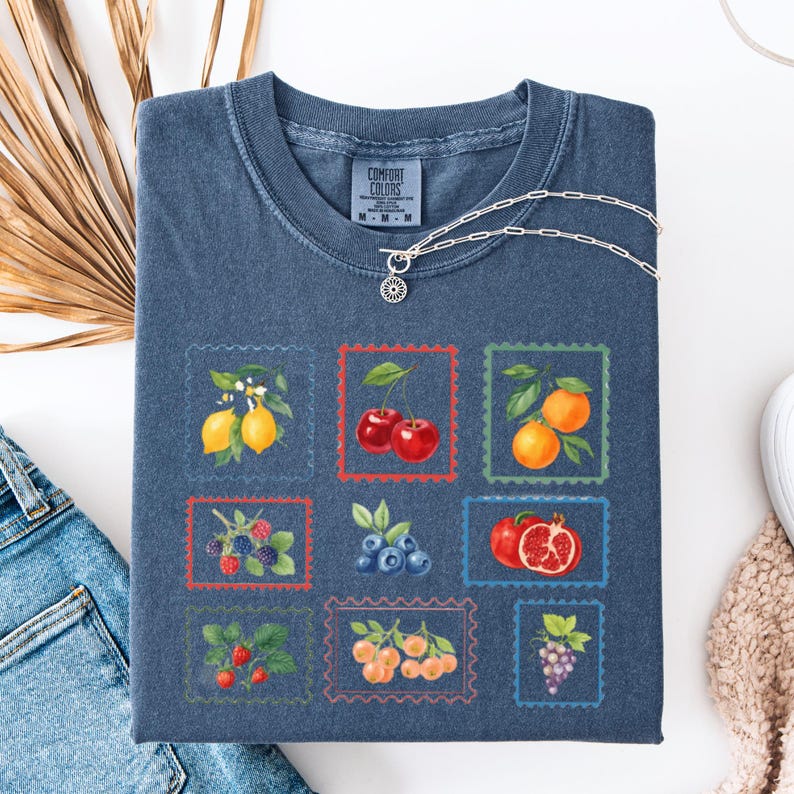 K&ouml;nnte beinhalten: Ein blaues Comfort Colors T-Shirt mit einem Raster von Obstillustrationen in Briefmarkenrahmen. Die Fr&uuml;chte umfassen Zitronen, Kirschen, Orangen, Brombeeren, Blaubeeren, Granat&auml;pfel, Erdbeeren, Pfirsiche und Trauben. Eine silberne Halskette liegt auf dem Shirt.