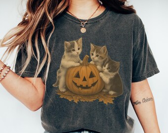 May include: Dark gray t-shirt featuring a Halloween design. The graphic shows three kittens gathered around a carved orange pumpkin. The pumpkin sits on a bed of autumn leaves. The kittens are in shades of gray, brown, and white.
