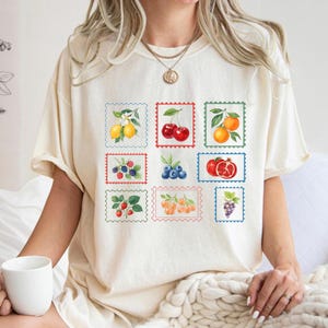K&ouml;nnte beinhalten: Cremefarbenes T-Shirt mit neun farbenfrohen Fruchtillustrationen in Briefmarkendesigns. Die Briefmarken zeigen Zitronen, Kirschen, Orangen, Brombeeren, Blaubeeren, einen Granatapfel, Erdbeeren, Aprikosen und Trauben. Das Shirt hat eine lockere Passform.