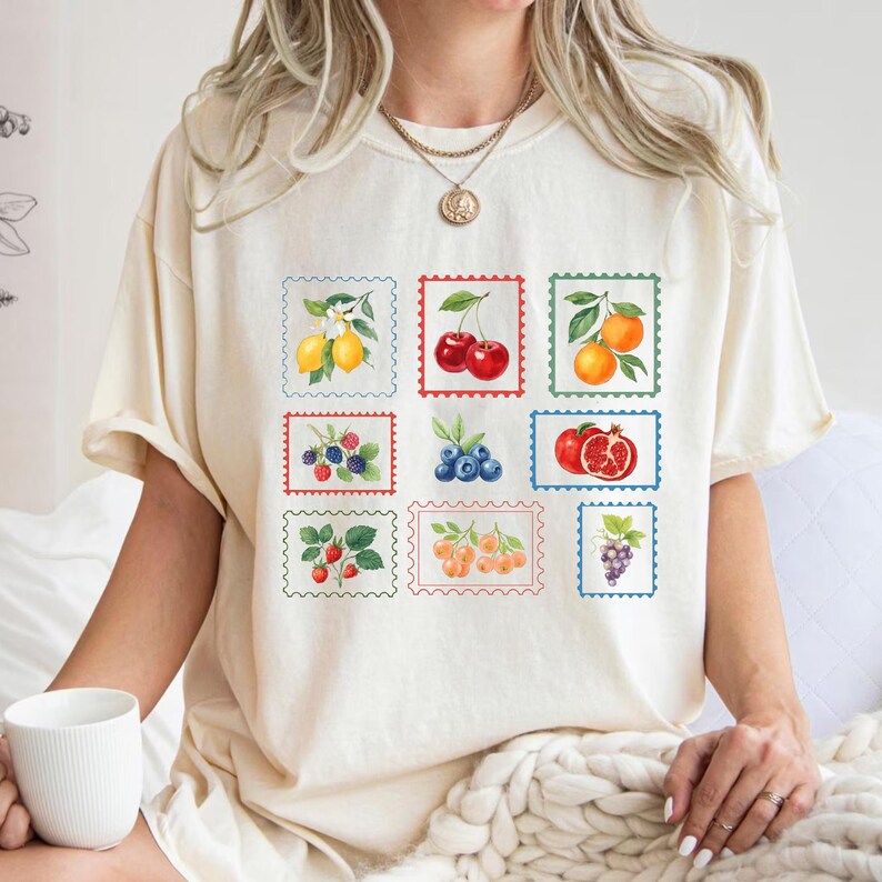 K&ouml;nnte beinhalten: Cremefarbenes T-Shirt mit neun farbenfrohen Fruchtillustrationen in Briefmarkendesigns. Die Briefmarken zeigen Zitronen, Kirschen, Orangen, Brombeeren, Blaubeeren, einen Granatapfel, Erdbeeren, Aprikosen und Trauben. Das Shirt hat eine lockere Passform.