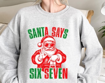 May include: A light gray sweatshirt with the text "SANTA SAYS" in green above a red illustration of Santa Claus wearing sunglasses and making a peace sign. The text "SIX SEVEN" is below the Santa illustration, in red and green.