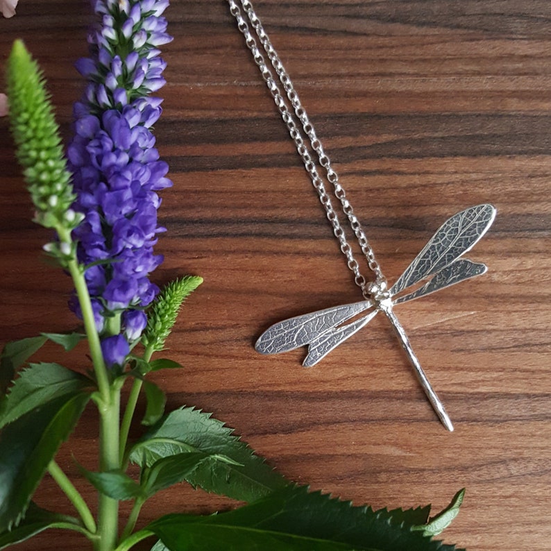 silver dragonfly necklace lying on a wooden background next to a purple flower. The wings are textured with the imprint of a leaf and the necklace chain is attached through a loop at the back of the head of the dragonfly