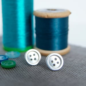 May include: Two silver buttons with four holes each, against a gray fabric background. The buttons are in focus, while two spools of teal thread are out of focus in the background.