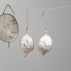 May include: Silver leaf-shaped drop earrings with a textured finish and three small, raised circles. The earrings are suspended from long, curved ear wires. A dried, round seed pod is also visible, suspended on a thin wire, adding a natural element.